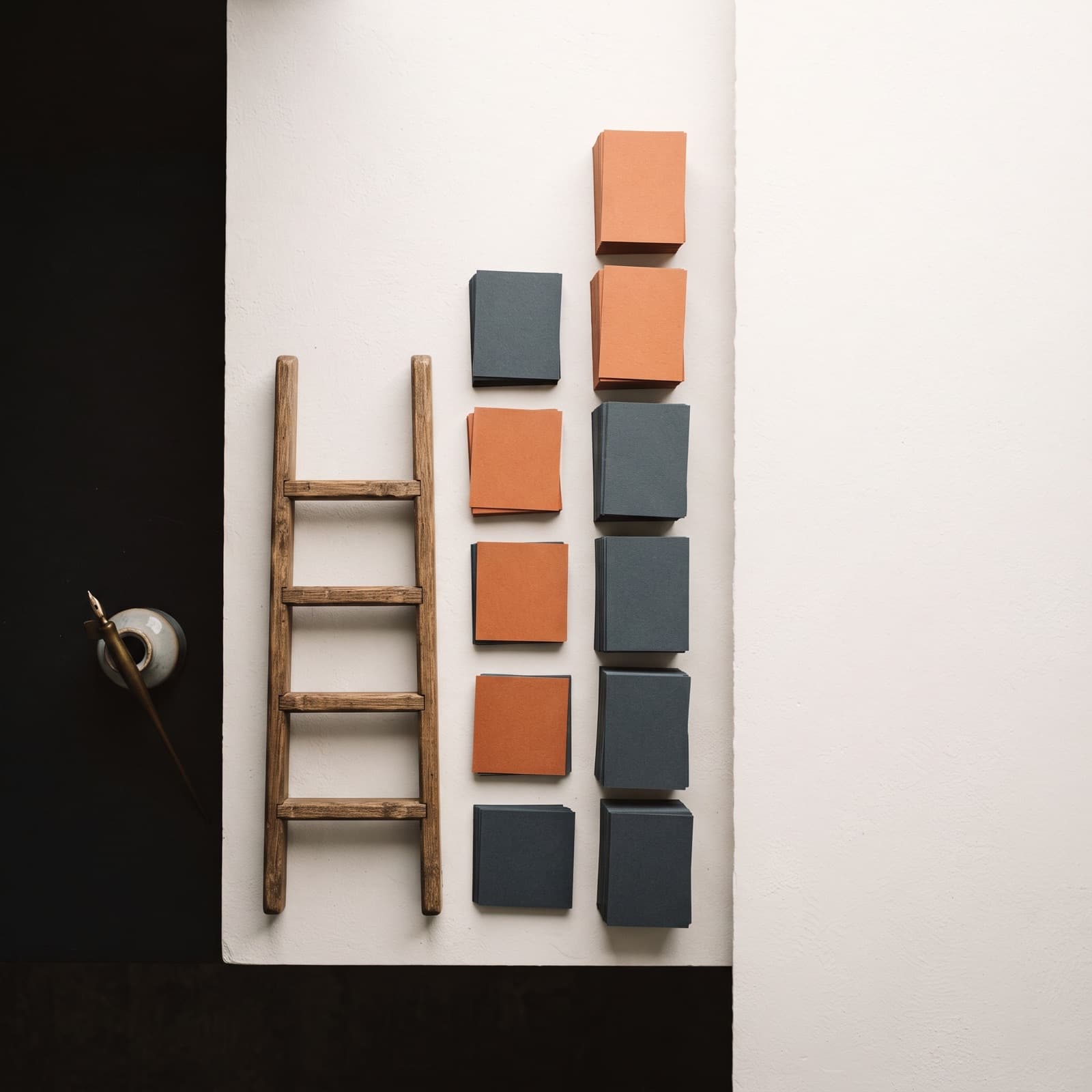 Editorial overhead scene of a four-rung wooden ladder motif on a warm white studio surface, with ascending stacks of printed ad frame cards beside each rung, a few cards tinted burnt orange.