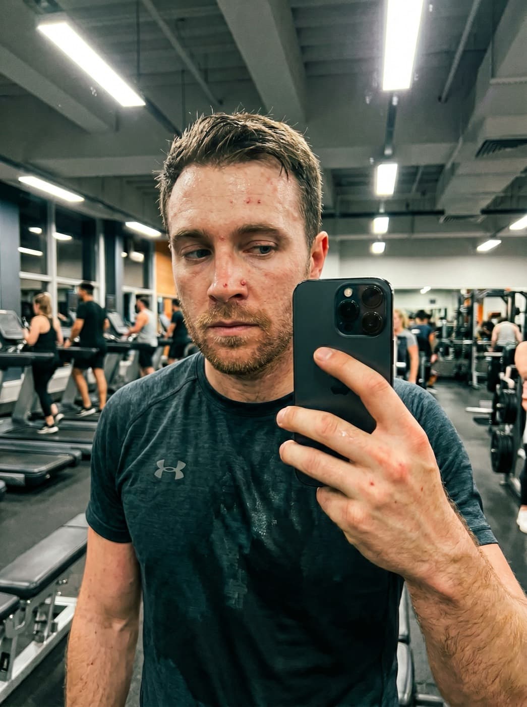 AI UGC creator post-workout mirror selfie in a busy gym, sweat on the brow, Under Armour tee