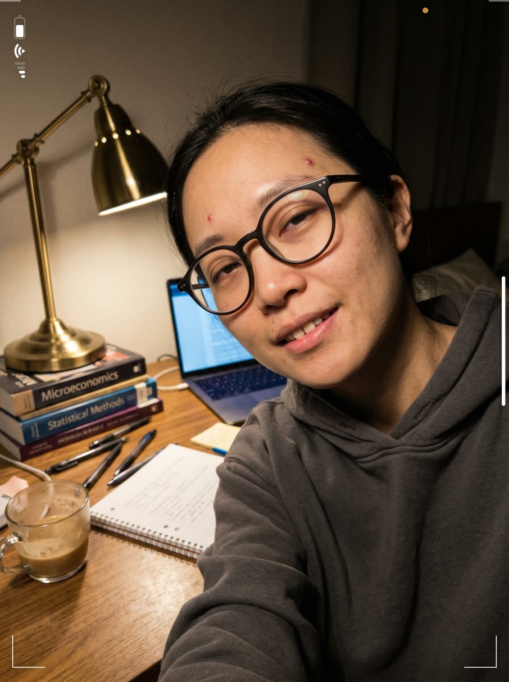 AI UGC creator selfie at a desk with brass lamp, latte and economics textbooks, late-night study vibe