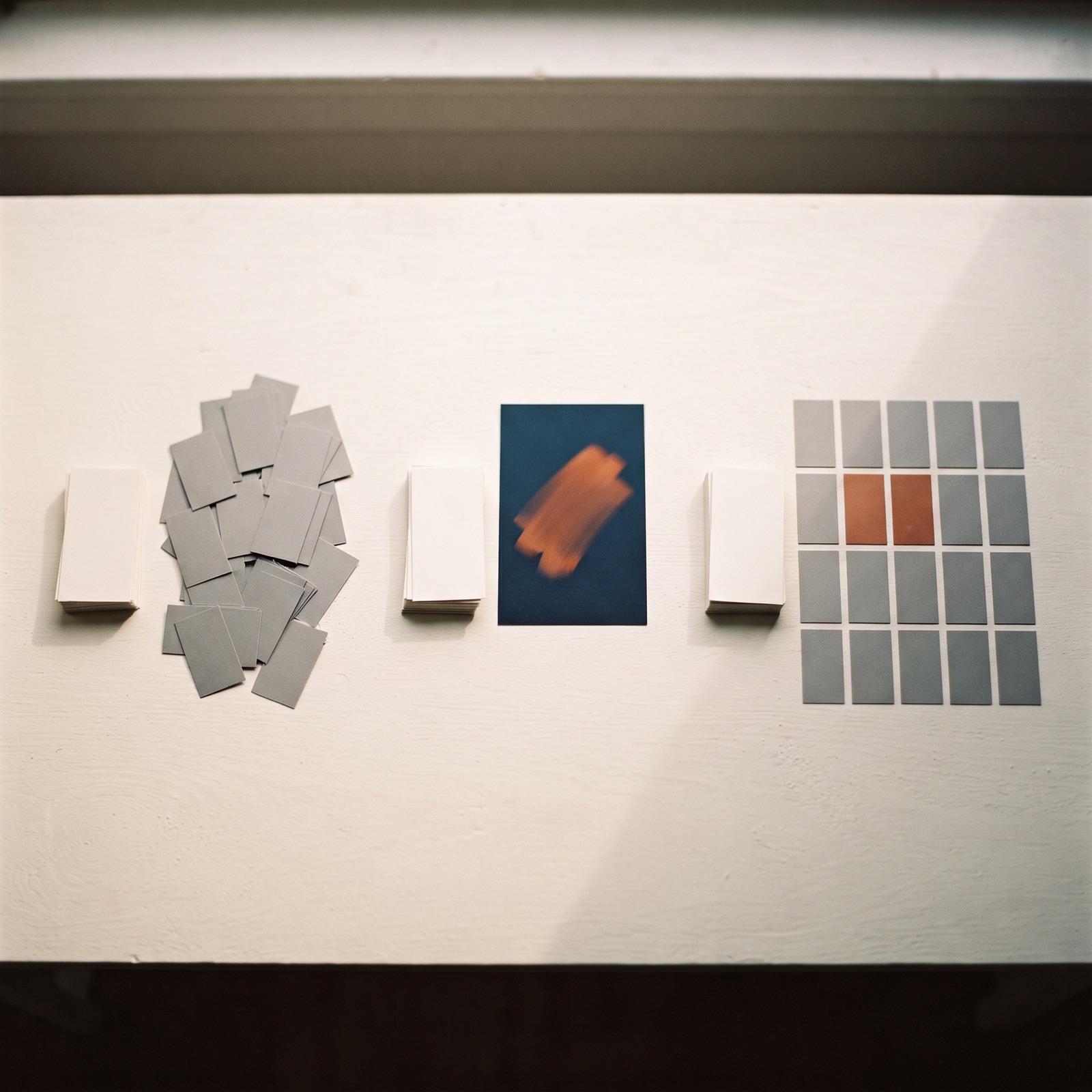 Overhead editorial scene of three groups of printed ad frame cards of visibly different sizes on a warm white desk, each preceded by an identical stack of blank paper slips, with two cards in the largest group tinted burnt orange.