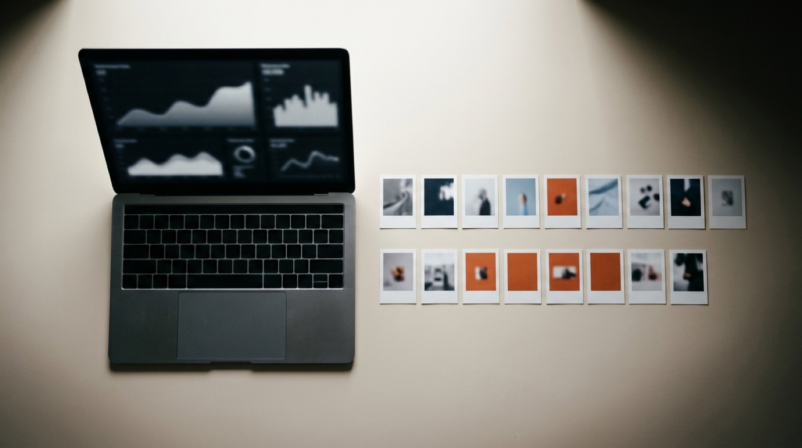 Overhead editorial scene of an open laptop on a warm white desk showing softly blurred rising and falling performance curves, beside two sorted rows of small printed ad frame cards with a few tinted burnt orange.