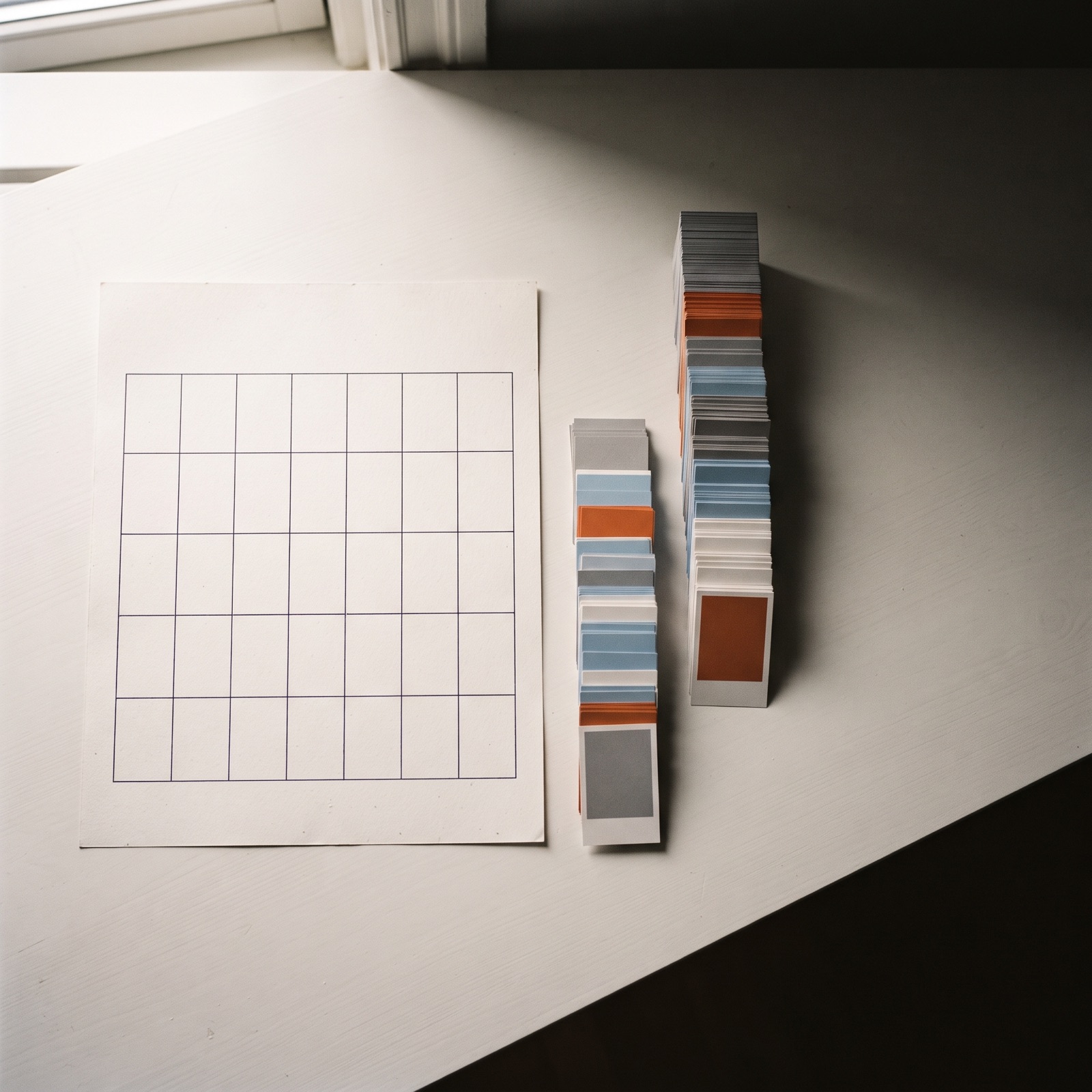 Overhead editorial scene of a wall calendar page beside four ascending stacks of printed ad frame cards on a warm white desk, a few cards tinted burnt orange among grey ones.