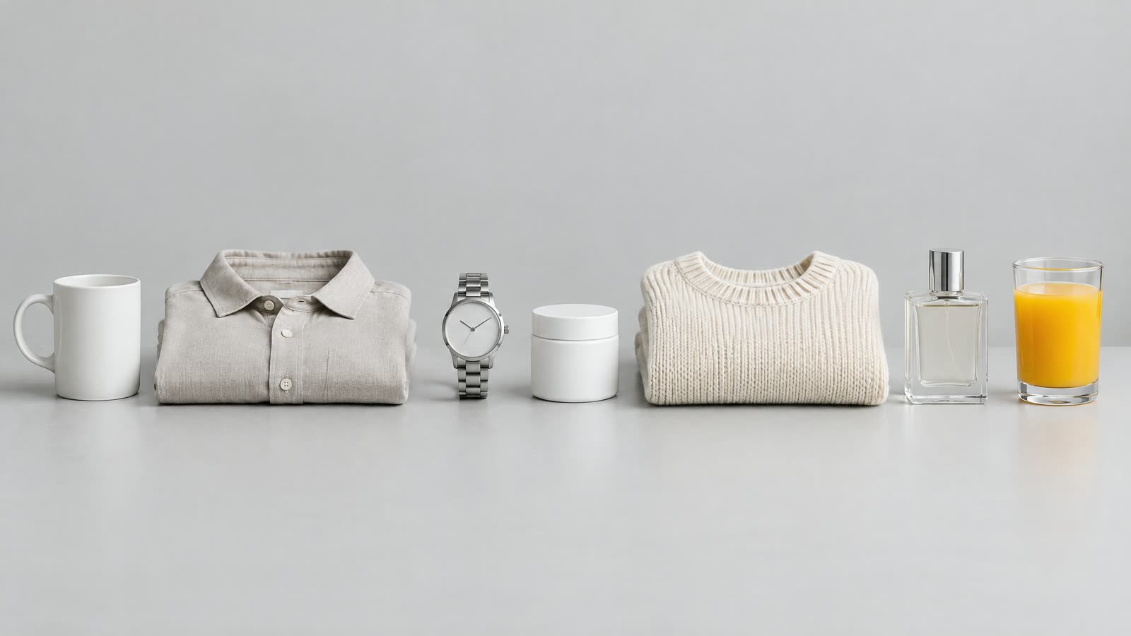 Syv produkter på række på et gråt studiebord: et mat keramikkrus, en foldet hørskjorte, et stålur, et hudplejeglas med blank etiket, en strikket sweater, en parfumeflaske i klart glas og et glas appelsinjuice