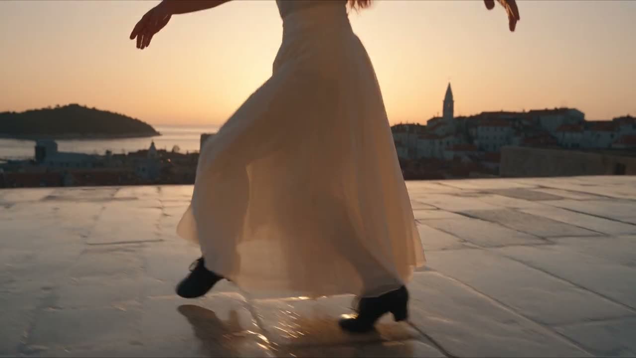 AI-generated cinematic fashion film: a woman in a flowing dress dancing on a Mediterranean rooftop terrace at sunset over a coastal old town.