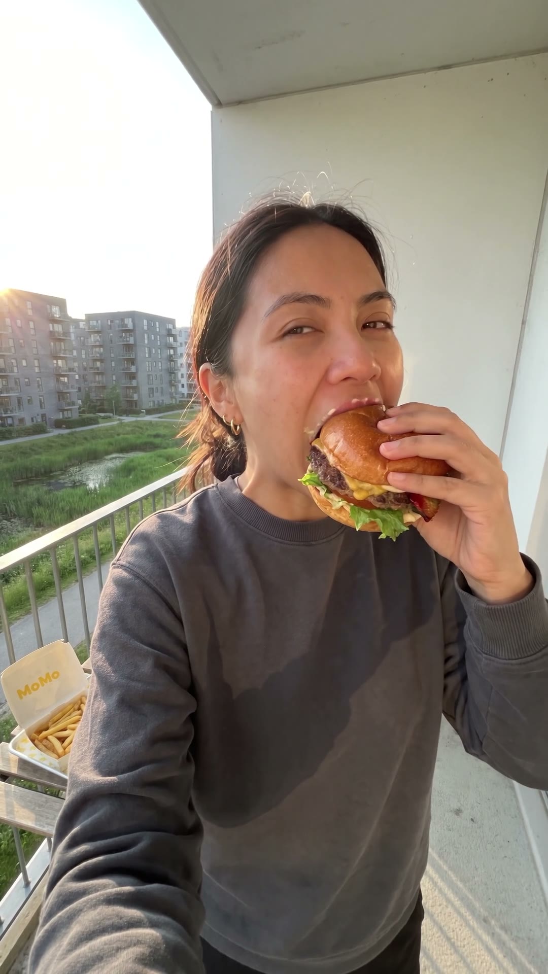 A woman takes the first bite of a MoMo burger on a balcony at golden hour, fries and takeaway bag beside her